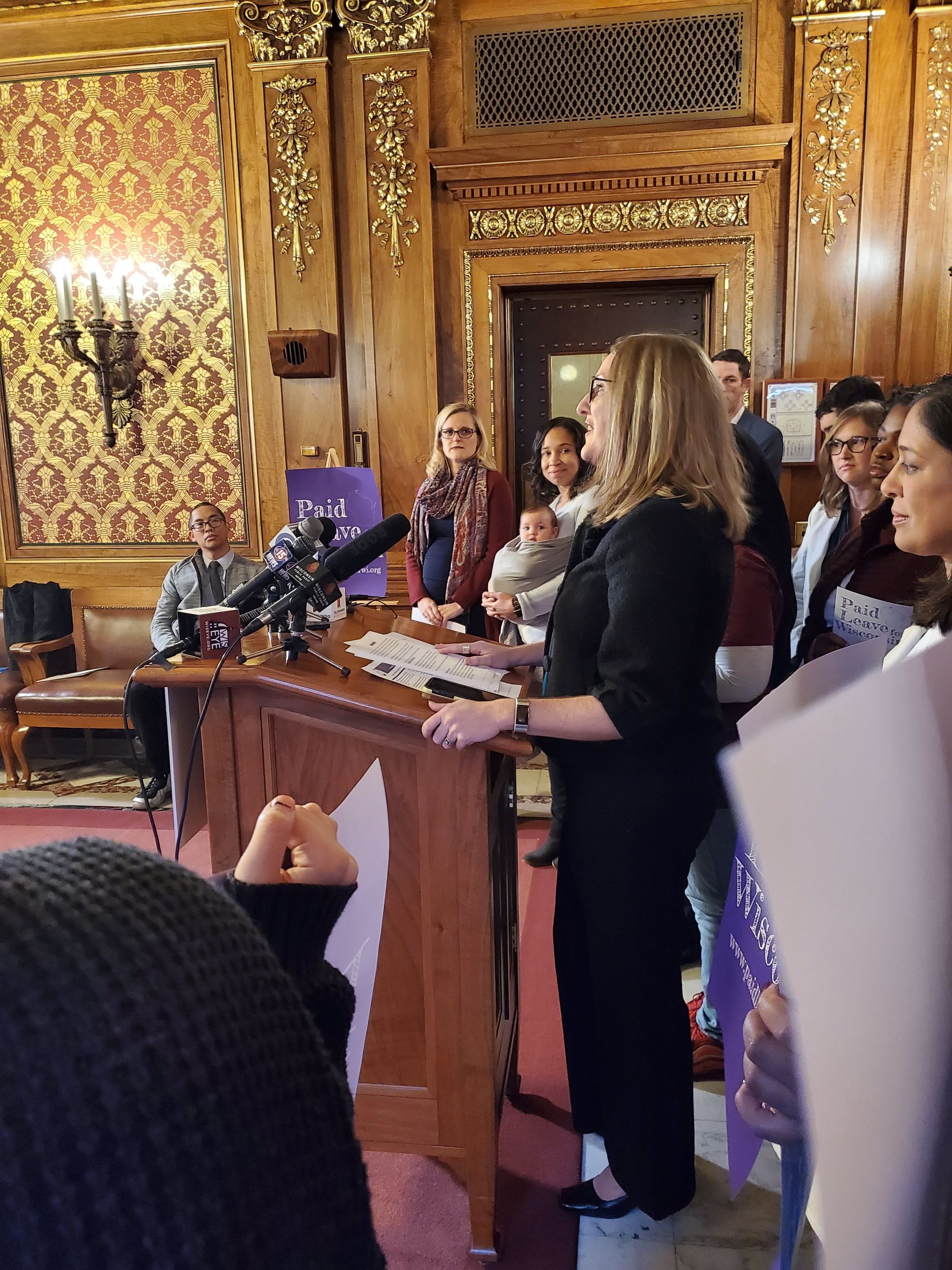 Image of a light skinned woman with light brown hair and dark glasses speaking at a wood podium. There are a variety of people standing around her, some with purple signs that say Paid Leave Wisconsin.       . 